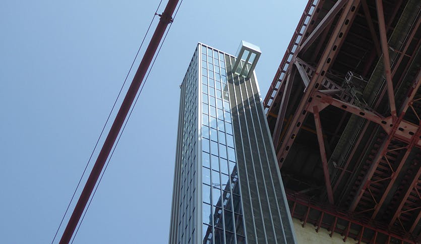 A tall, glass-clad building with a protruding glass box near the top, adjacent to a large, red metal structure.