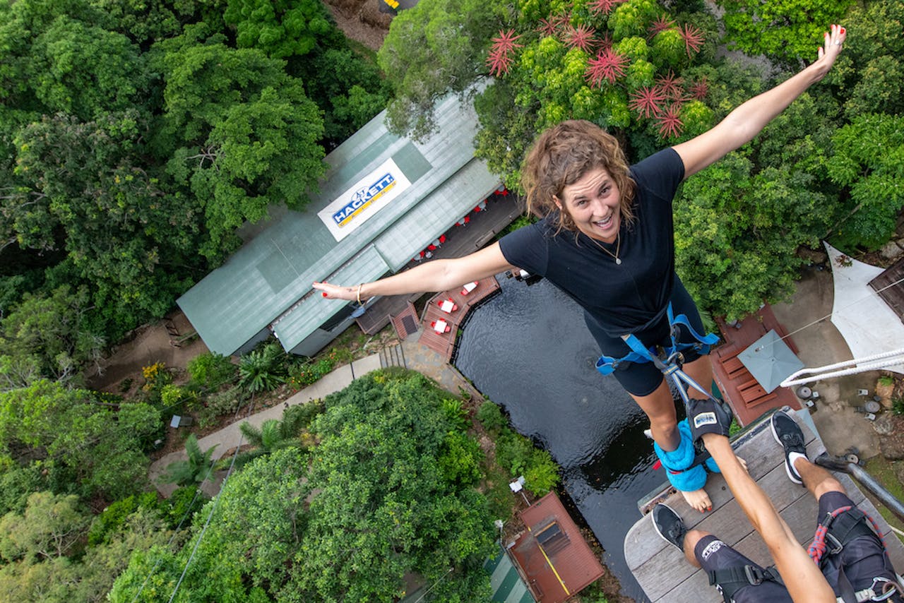 A woman in a harness leans forward with outstretched arms, preparing to bungee jump from a high platform surrounded by greenery.