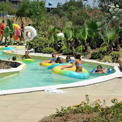 Barn och vuxna kopplar av på färgglada uppblåsbara rör i en lat flod vid en vattenpark omgiven av frodig vegetation och vägar.