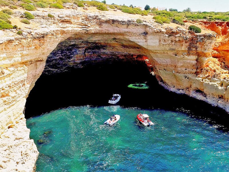 Three small boats floating in clear blue water inside a large sea cave with rugged, rocky walls and a green bush-covered top.