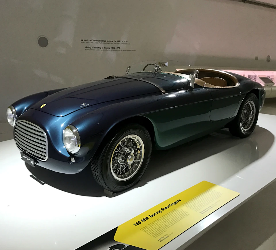 A dark blue vintage sports car with exposed wheels displayed in a museum, accompanied by a yellow information plaque.