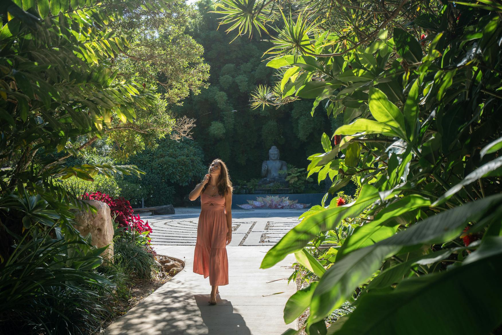 珊瑚色のドレスを着た女性が、仏像を背景に、緑豊かな緑に囲まれた庭園の小道を歩いています。