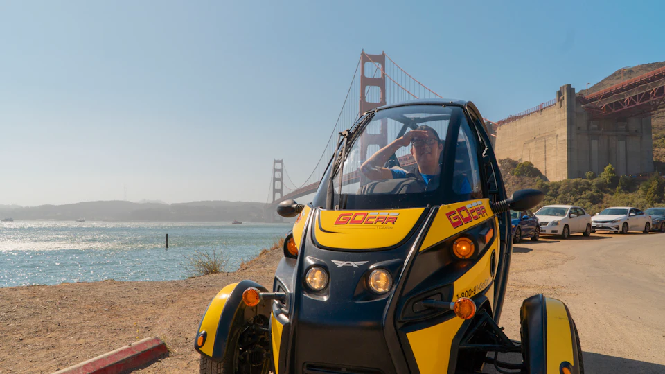 A person sits in a yellow and black GoCar with the Golden Gate Bridge visible in the background on a sunny day.
