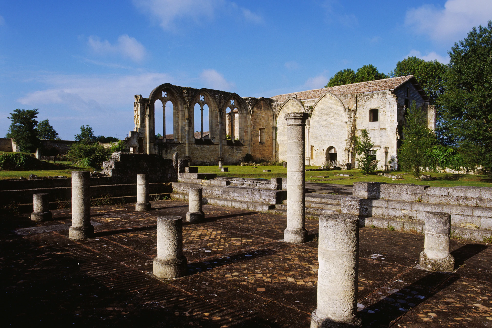Ruins of an old stone building with tall columns and arched windows under a clear blue sky. Trees in the background.
