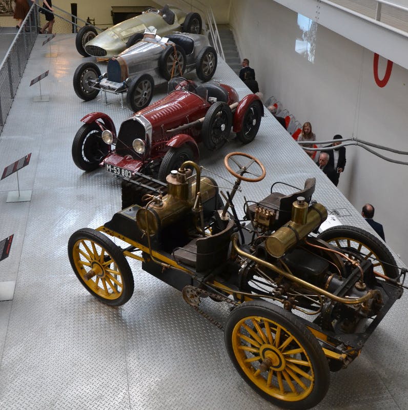 Four vintage cars displayed on a metal platform with a group of people observing them from below.