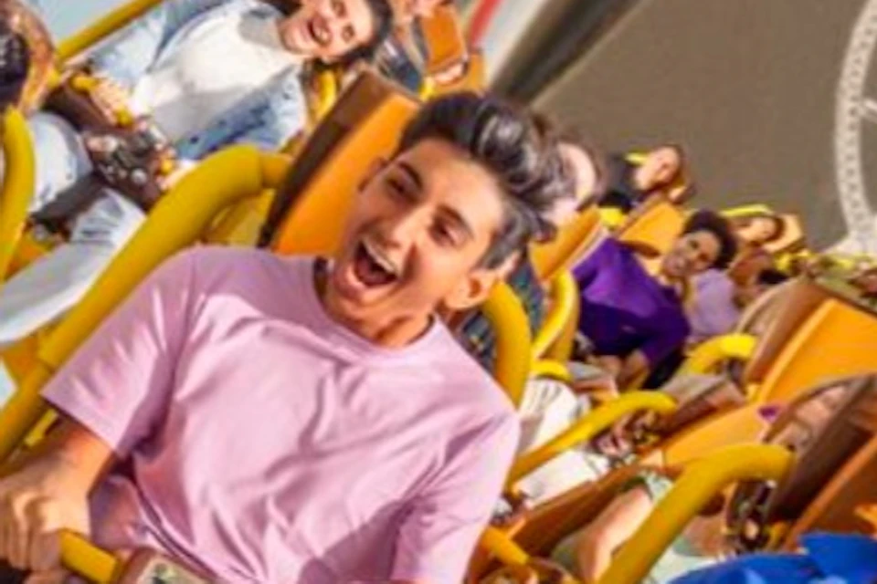 People riding a yellow roller coaster with visible tracks and a building in the background under a blue sky.