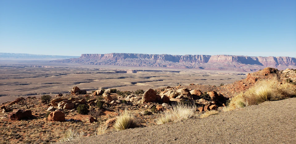 Expansive desert landscape with sparse vegetation and large rock formations in the foreground. Distant mesas under a clear blue sky.