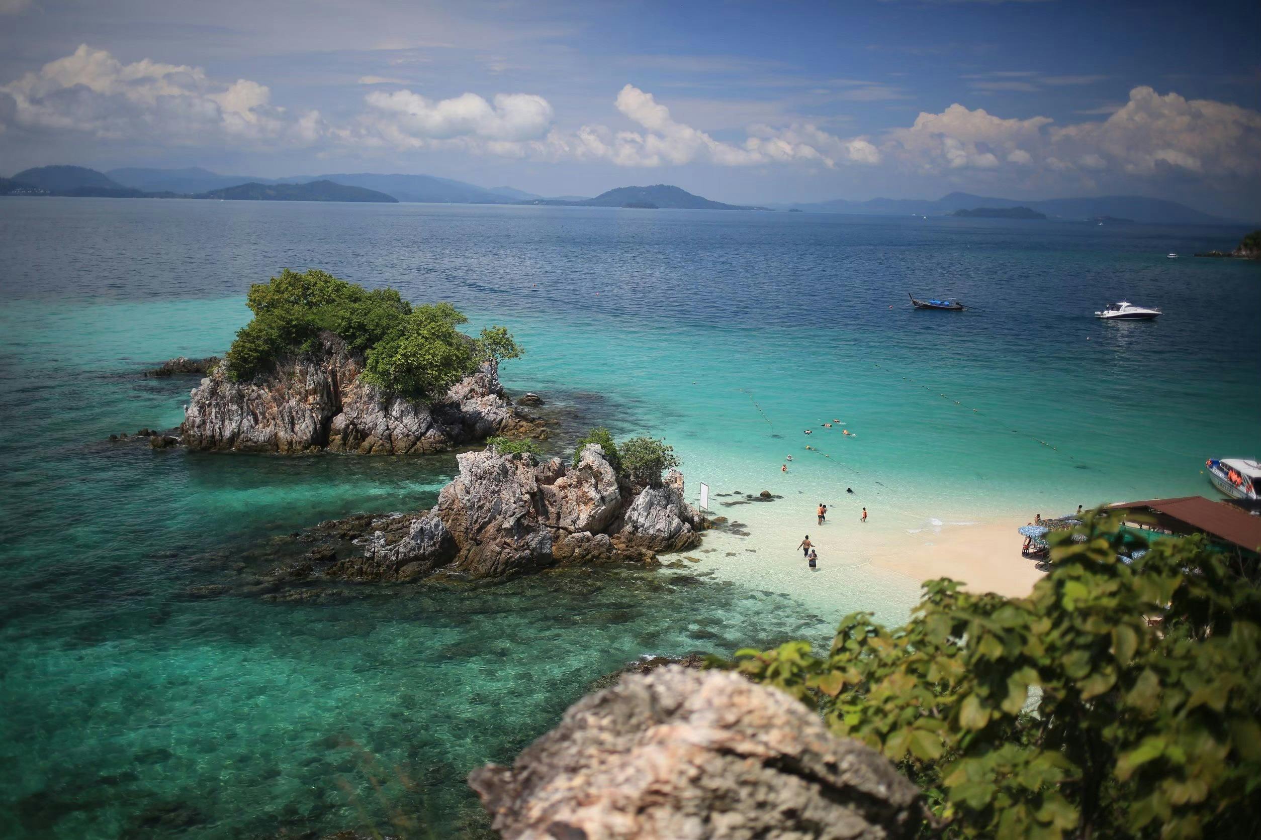 People enjoying a sandy beach with clear turquoise water, rocky outcrops, and a boat anchored nearby under a partly cloudy sky.