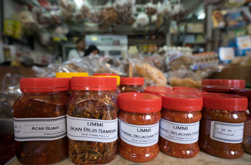 Rows of jars with red lids labeled "UMMI" on a market counter. Blurred shelves with various packaged goods in the background.