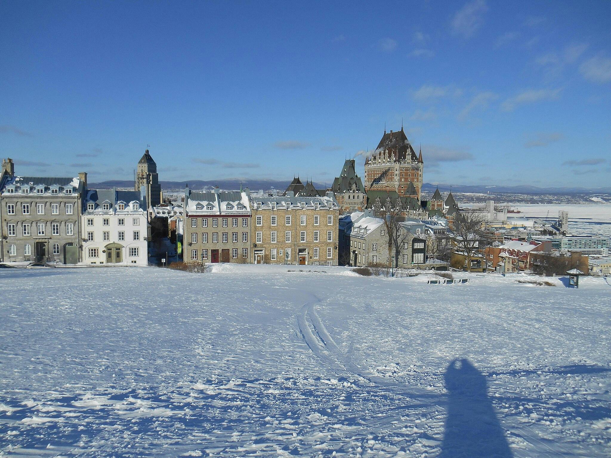 Snow-covered field with footprints leading to historic buildings, including a large chateau, under a clear blue sky.