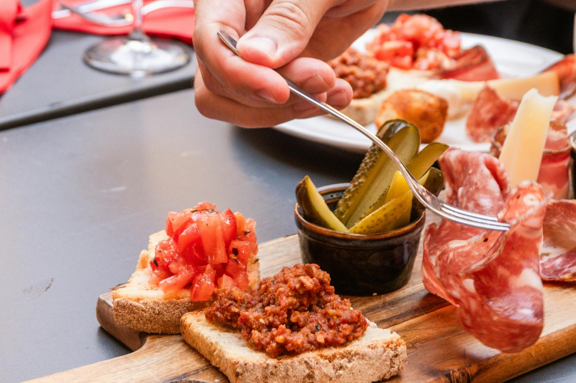 Hand using a fork to arrange toppings on bread slices, with diced tomatoes, minced meat, and pickles on a wooden platter.
