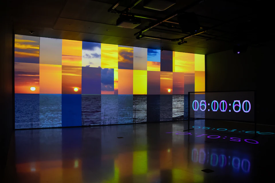 A modern art display featuring a segmented projection of a sunset over the ocean, with a digital clock reading 05:00.