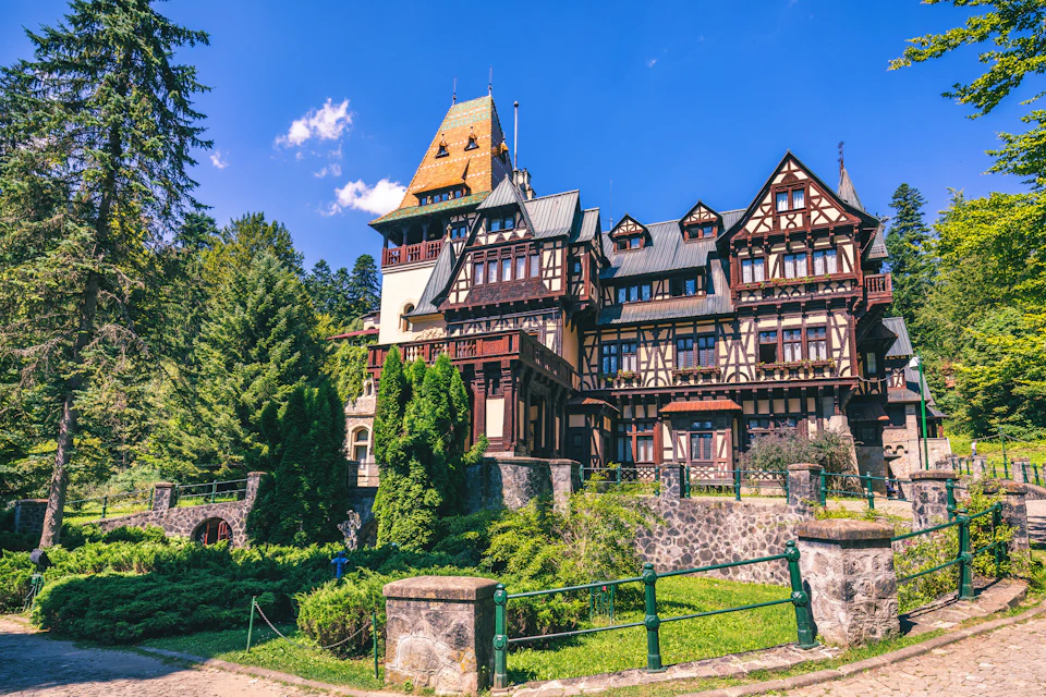 A large, ornate building with steep roofs and timber framing, surrounded by greenery and set against a clear blue sky.