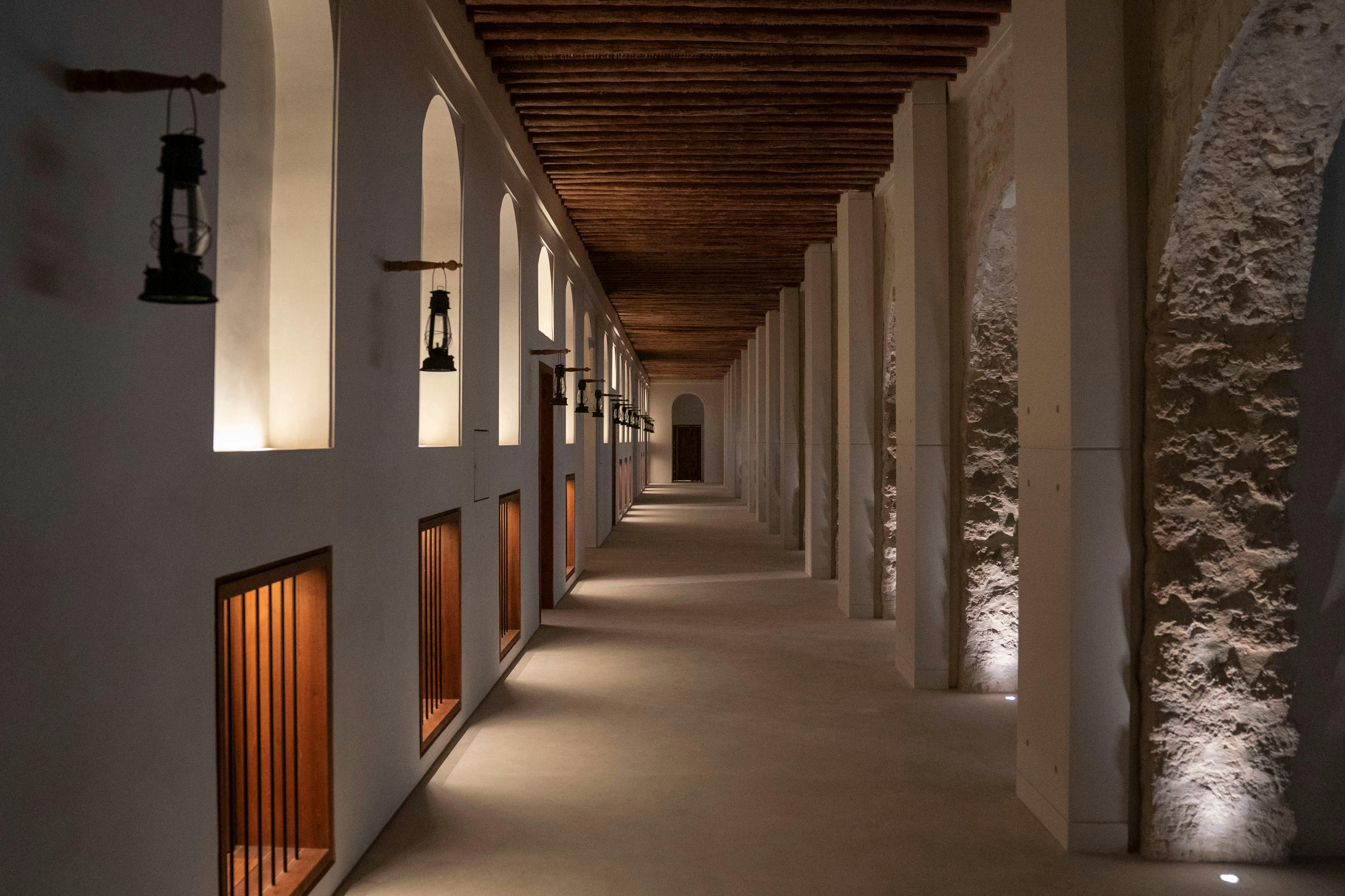 Long, dimly lit corridor with tall arches, wooden ceiling beams, and small lanterns hanging on the walls.