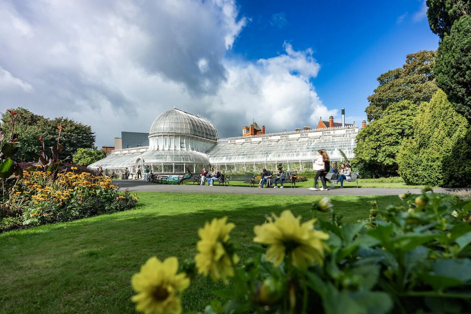 A botanical garden with a glass conservatory building, surrounded by green lawns, flowers, and people walking or sitting.