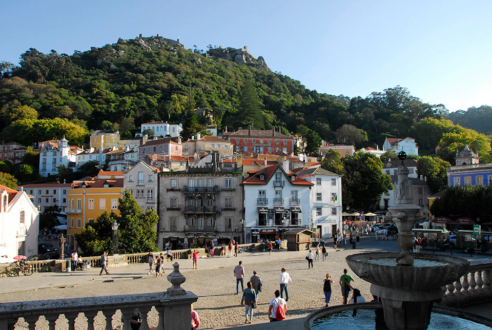 People walking in a plaza surrounded by colorful, historic buildings, with a green, forested hill in the background.