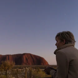 A person in a coat looks at a large rock formation under a twilight sky.