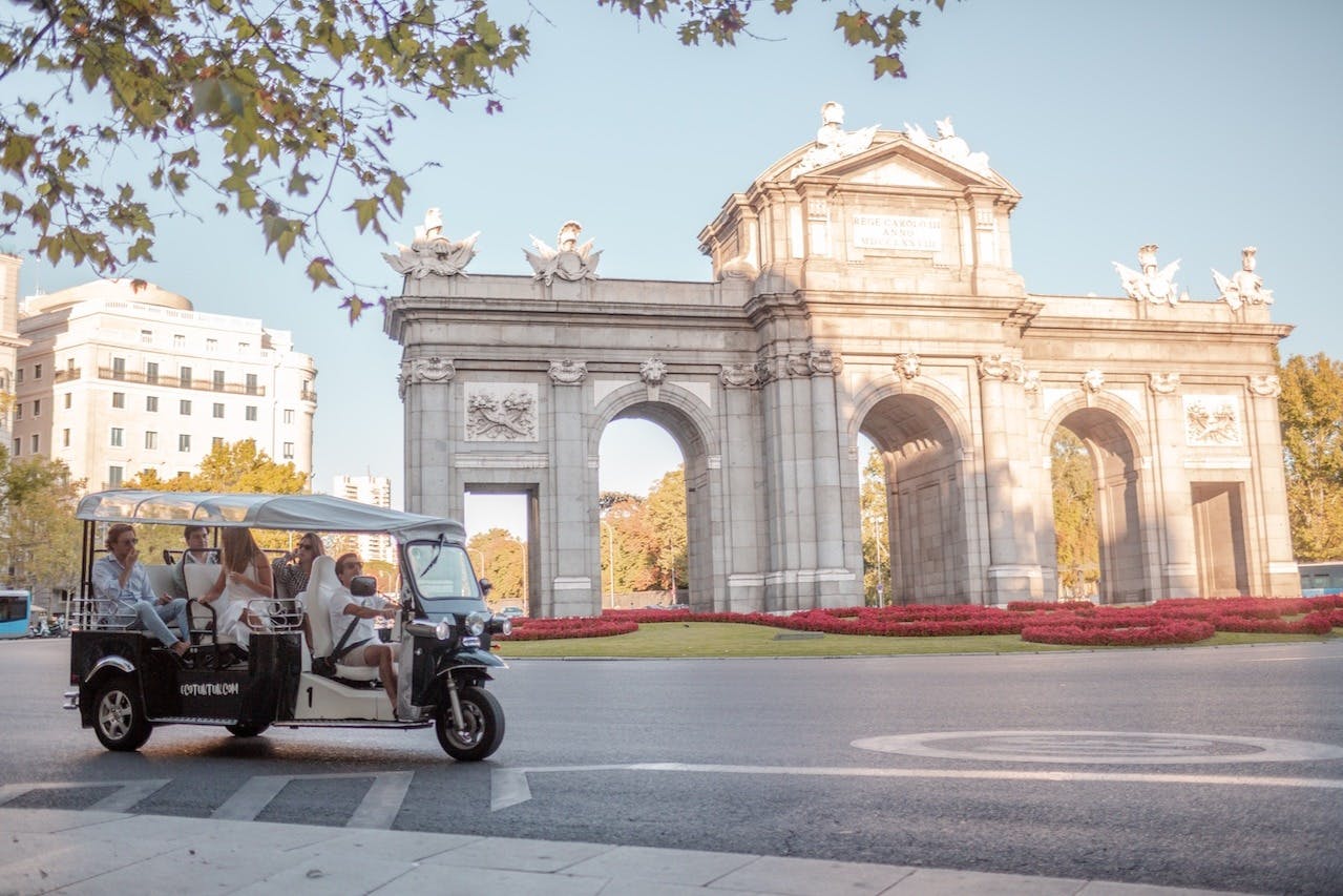 A tuk-tuk drives past the Puerta de Alcalá in Madrid, with trees and city buildings in the background.