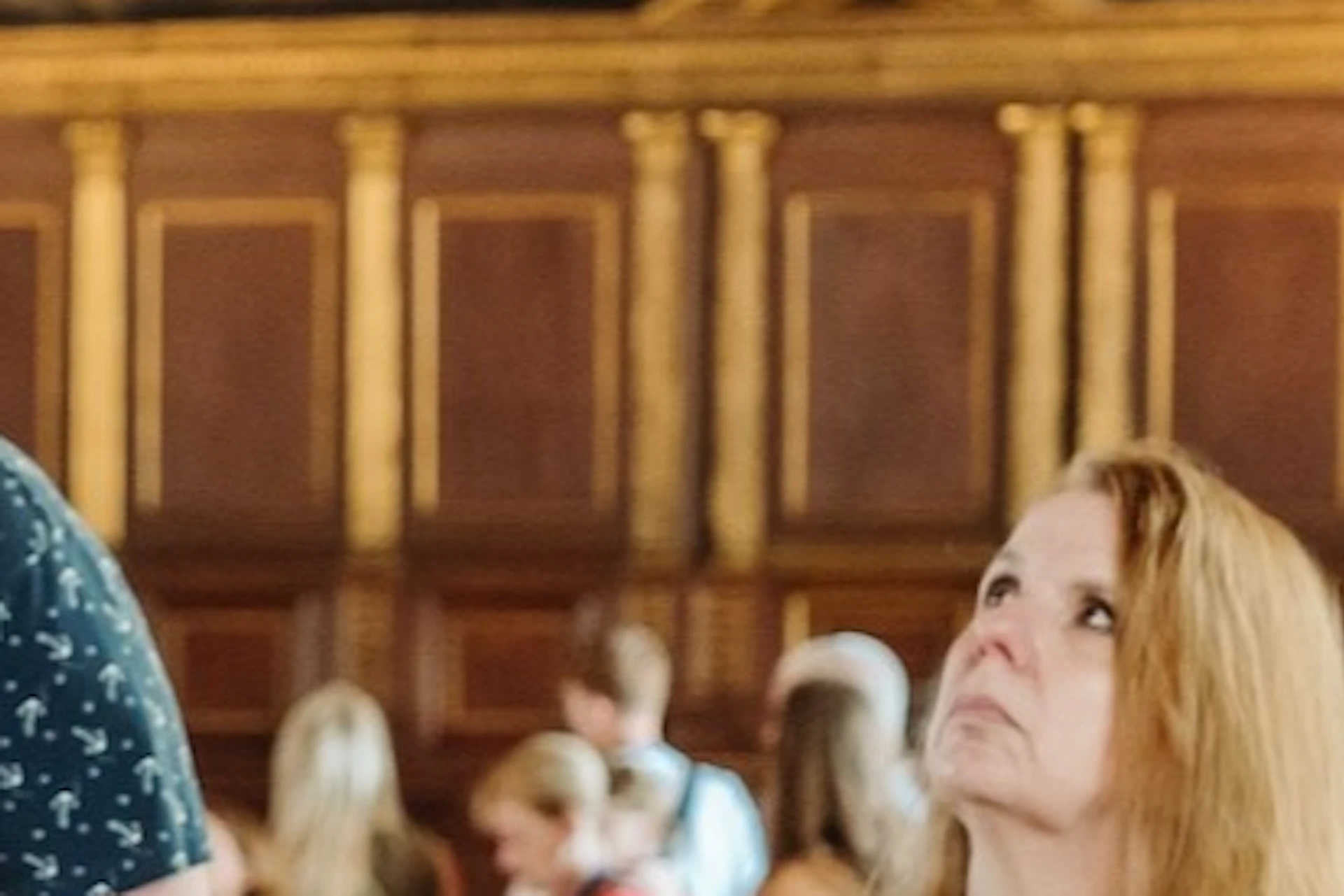 A guide gestures while explaining art to a group of visitors in an ornate museum hall with a large mural on the wall.
