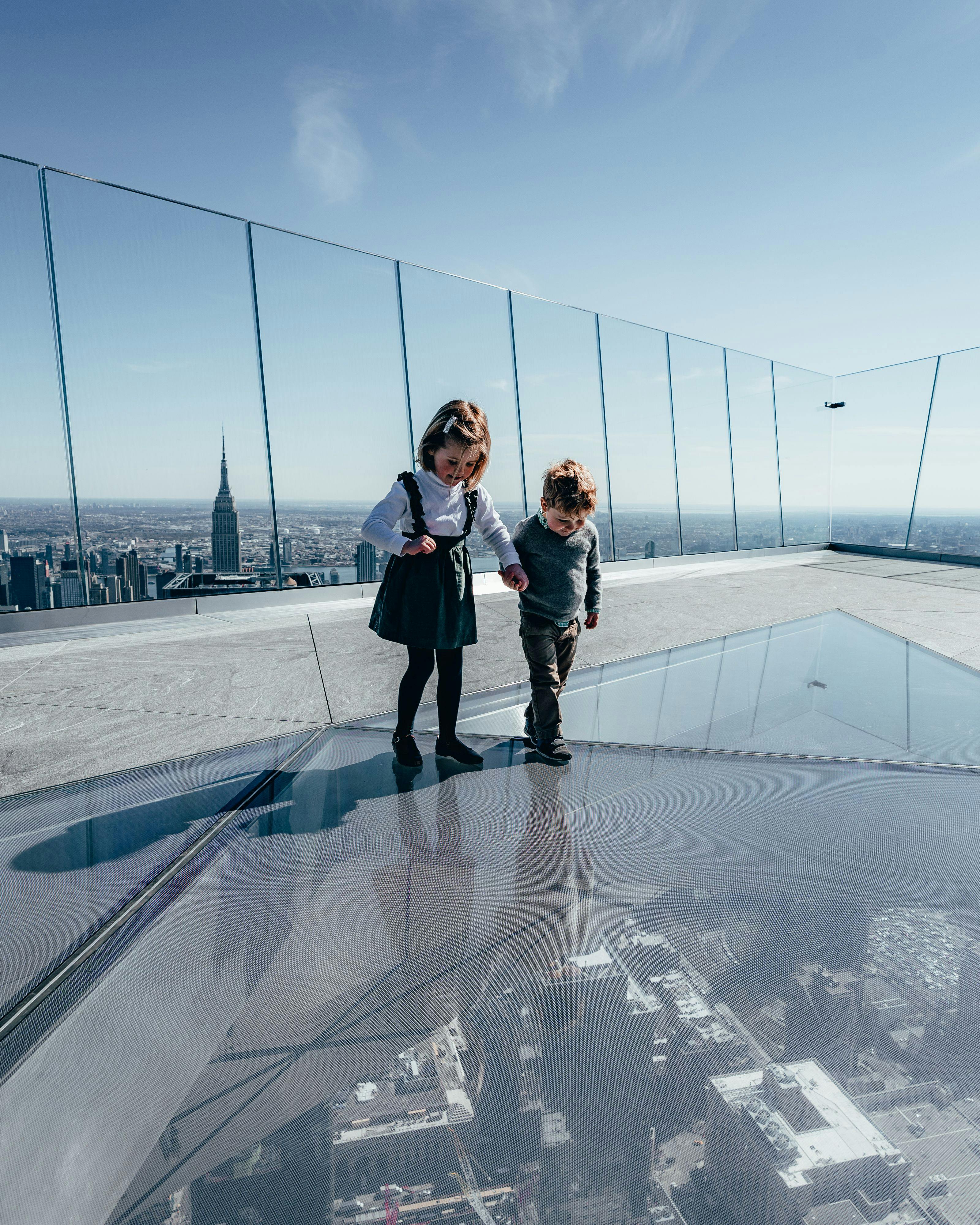 Two children holding hands stand on a glass floor at a rooftop observation deck, with a city skyline in the background.