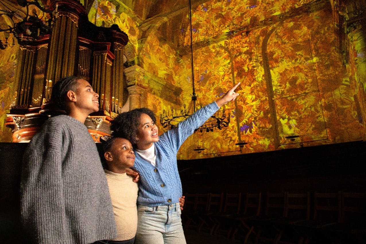 Τρία άτομα στέκονται μαζί, κοιτάζοντας προς τα πάνω με δέος μια πολύχρωμη, φωτιζόμενη οροφή σε ένα μεγαλοπρεπές εσωτερικό περιβάλλον.