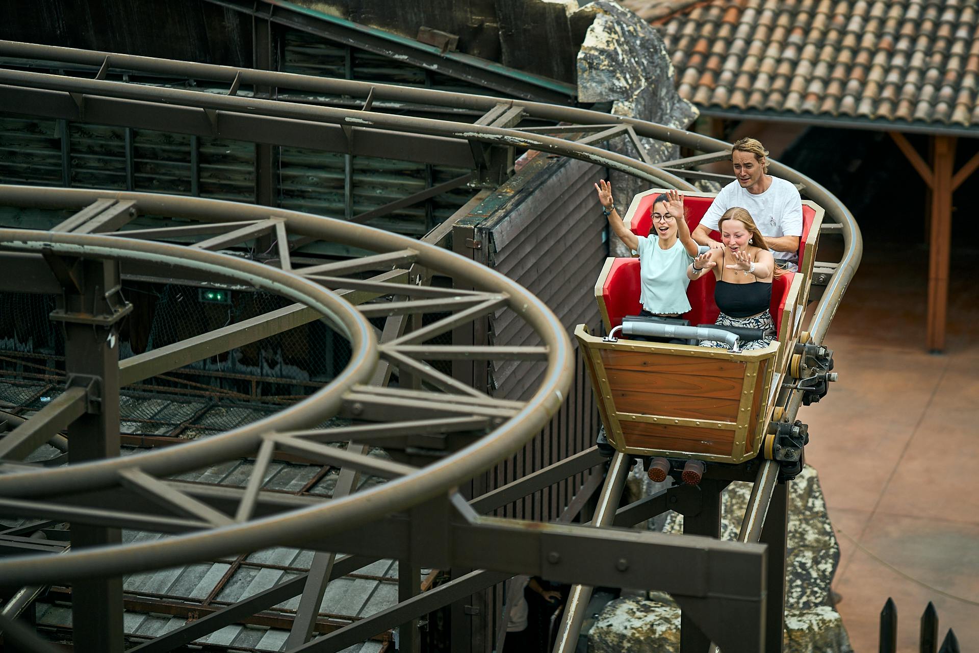 Tři lidé jezdí na horské dráze s výrazy vzrušení, míjejí skály a budovy.