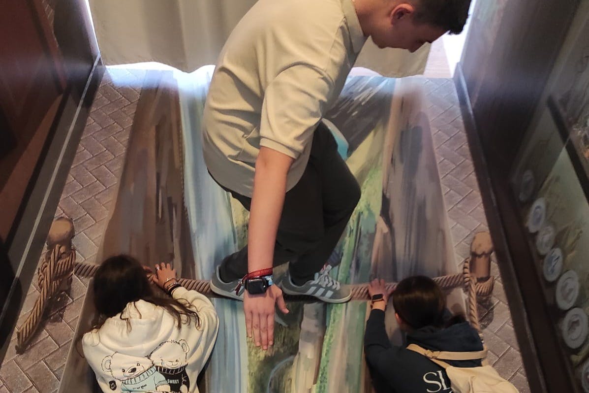 Tres persones interactuen amb una pintura de terra d'un pont de corda sobre un barranc, creant un efecte d'il·lusió 3D.