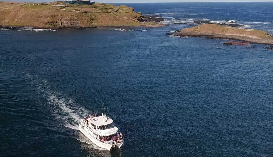 A boat filled with people sails on blue water near a rugged coastline with grassy hills and distant rocky formations.