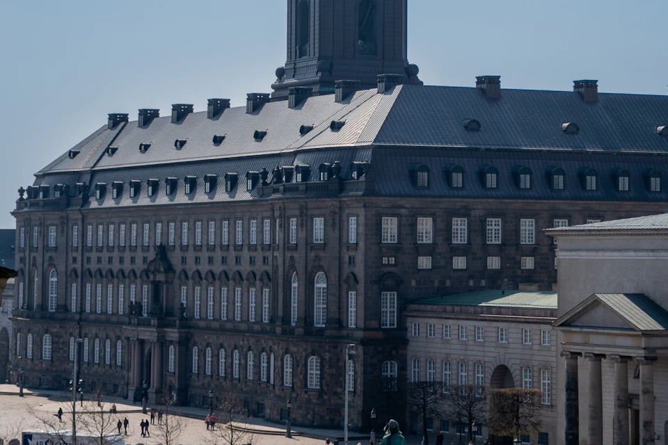 Large, historic building with numerous windows and a tall clock tower, people walking and gathering in a spacious courtyard below.