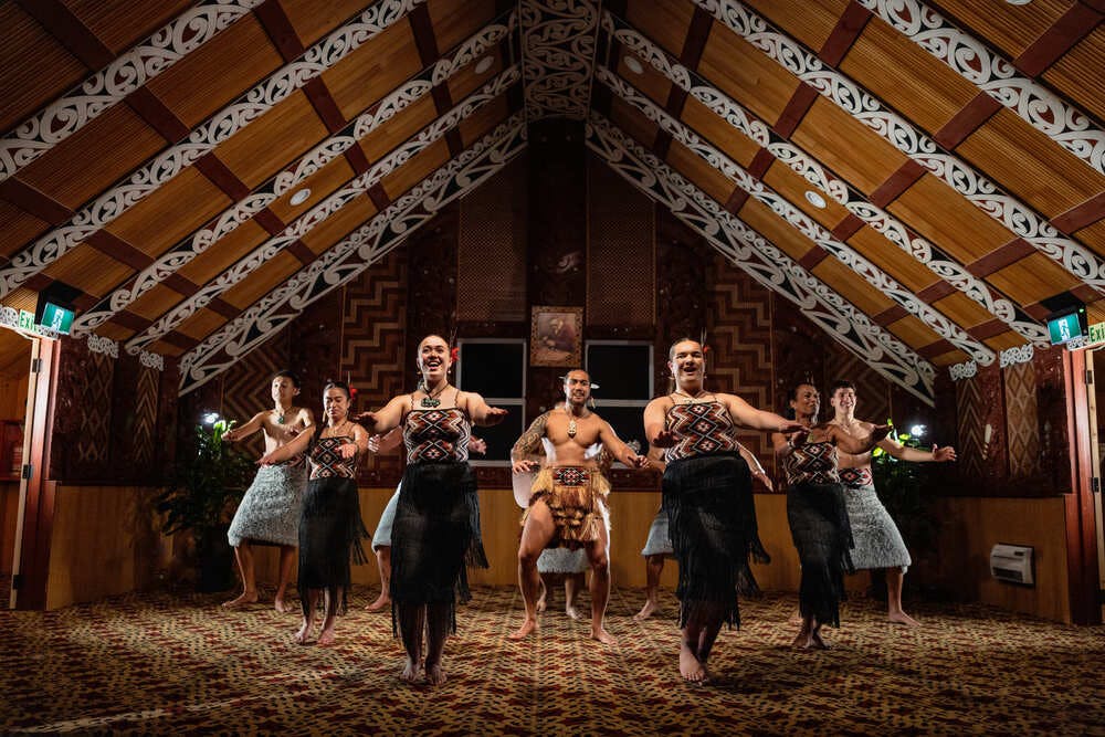 Performers in traditional attire dancing in a decorated wooden hall with intricate patterns.