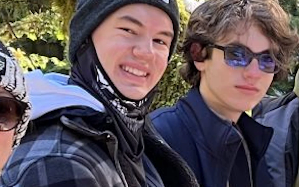 Four people stand on a wooden bridge in a forested area, dressed in jackets and hats, smiling at the camera.