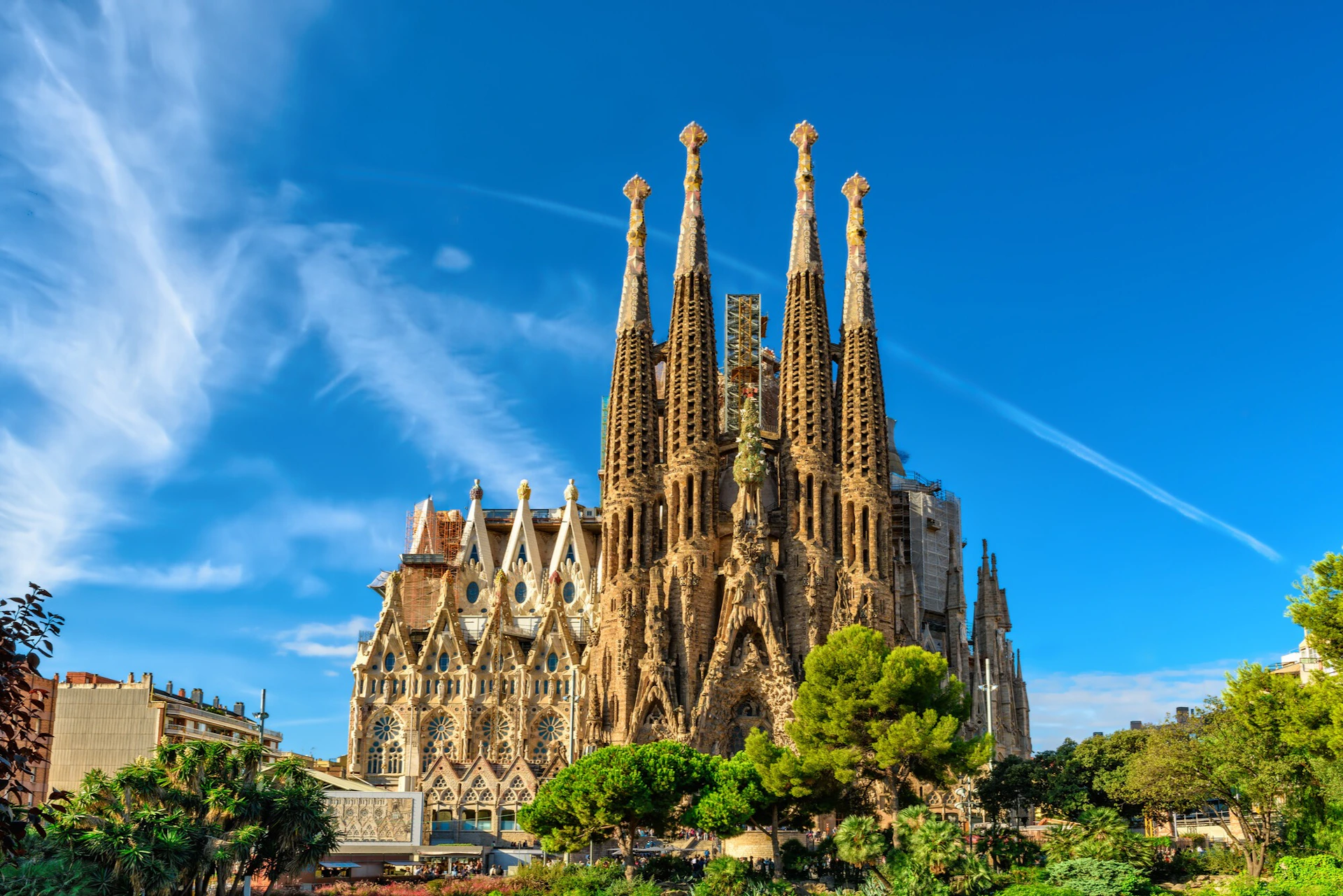 A large, ornate cathedral with multiple spires under a clear blue sky, surrounded by trees and city buildings.