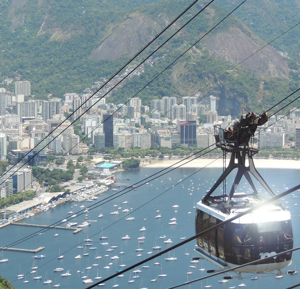 Cable car suspended over a bay with numerous boats, set against a backdrop of city buildings and green mountains.