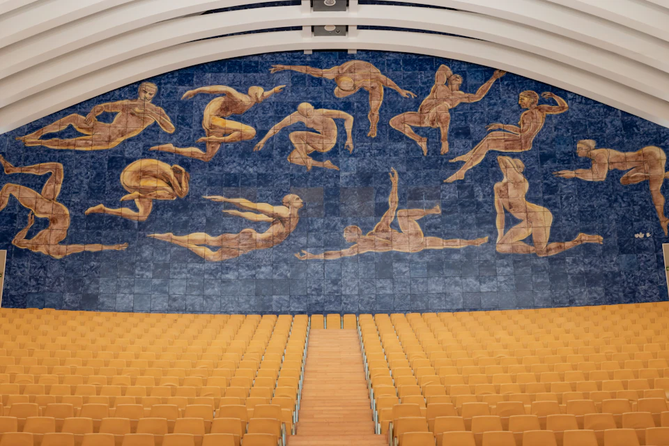 A lecture hall with rows of yellow seats and a large mural of floating human figures on a blue background.