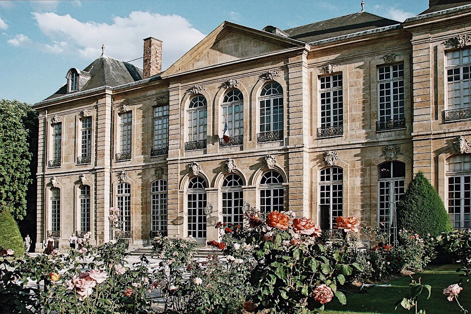 Historic building with arched windows and decorative stonework, surrounded by a blooming rose garden under a clear blue sky.