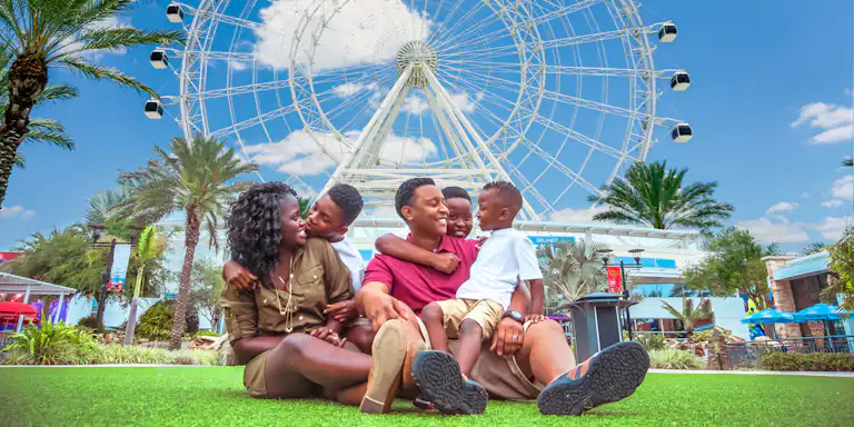 Eine fünfköpfige Familie sitzt lächelnd im Gras vor einem großen Riesenrad, mit Palmen und einem klaren blauen Himmel im Hintergrund.