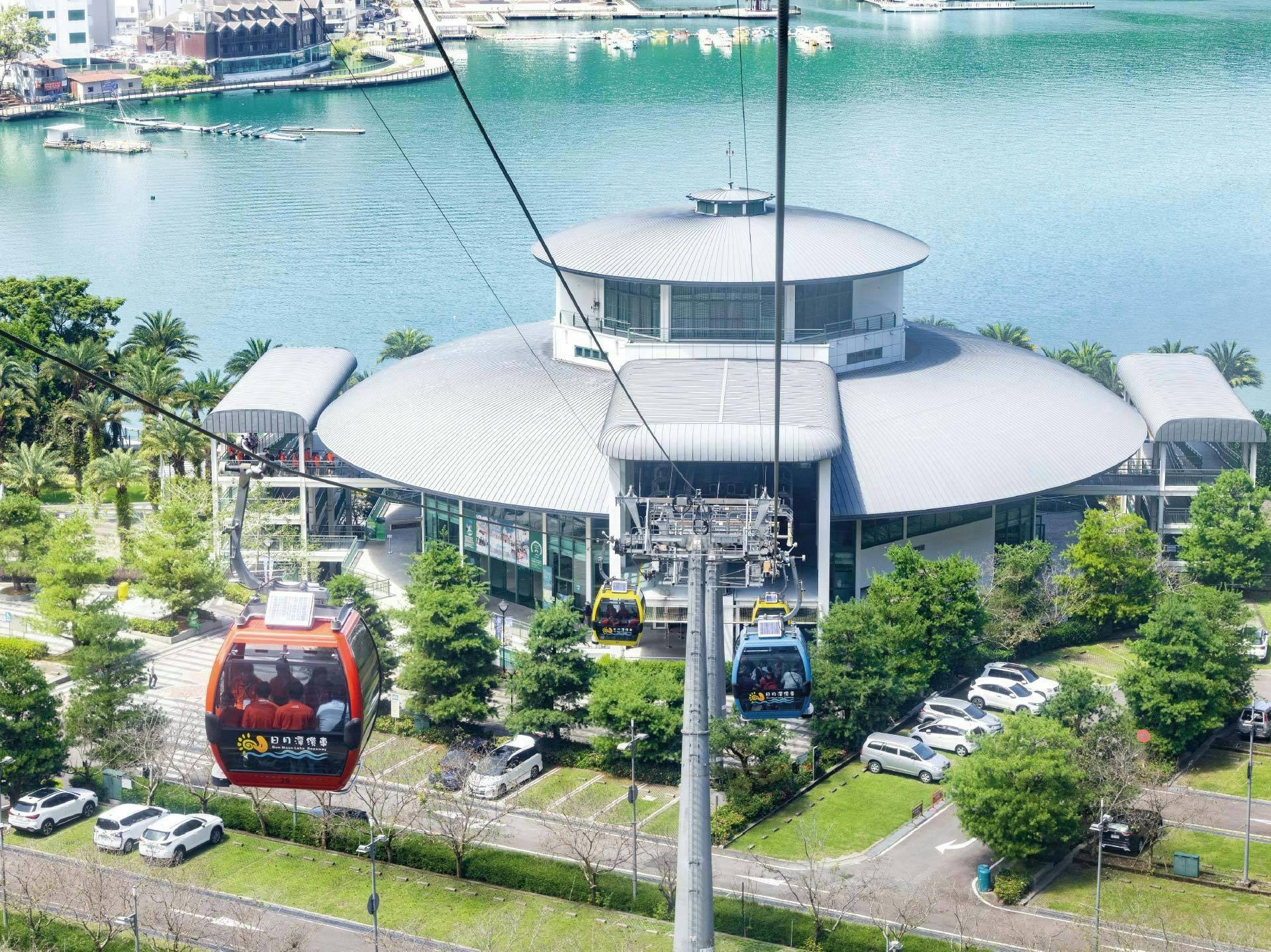 Cable cars moving over a scenic area with a round building, green trees, and a body of water in the background.