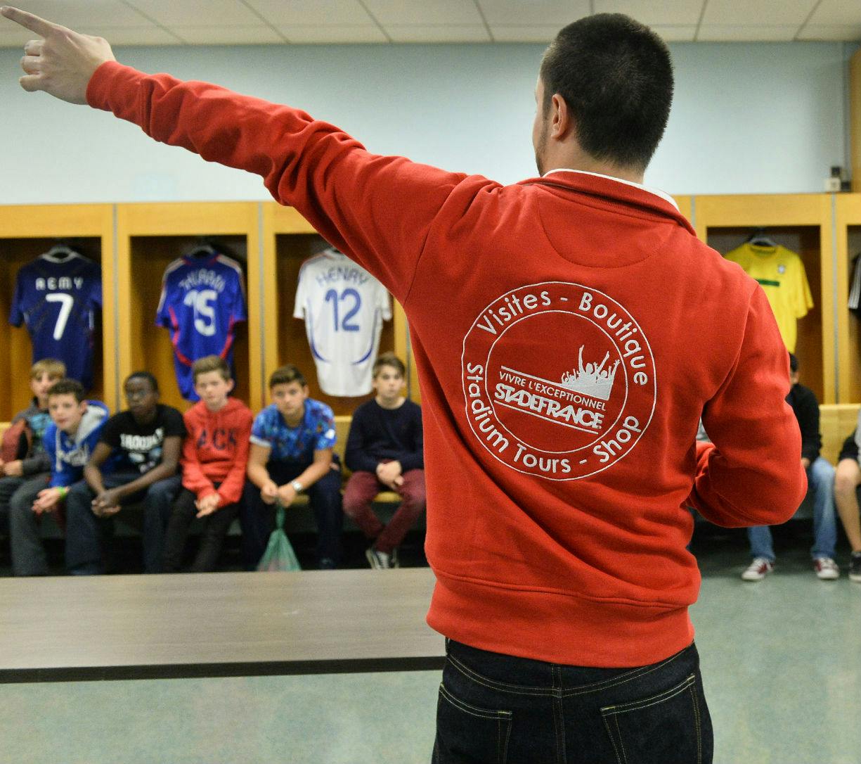 赤いジャケットを着た大人が、背後にジャージが掛けられたロッカールームに座っている若者のグループを見学しています。