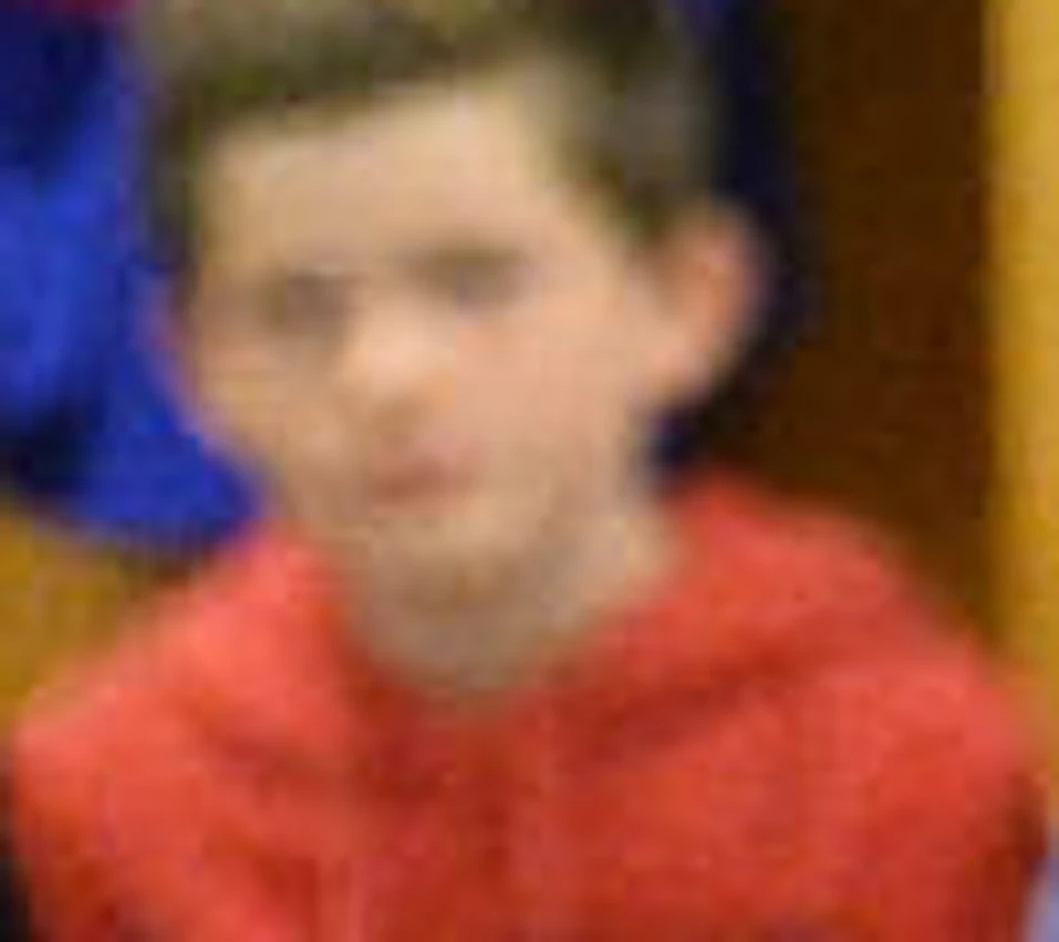An adult in a red jacket is giving a tour to a group of young people seated in a locker room with jerseys hanging behind them.