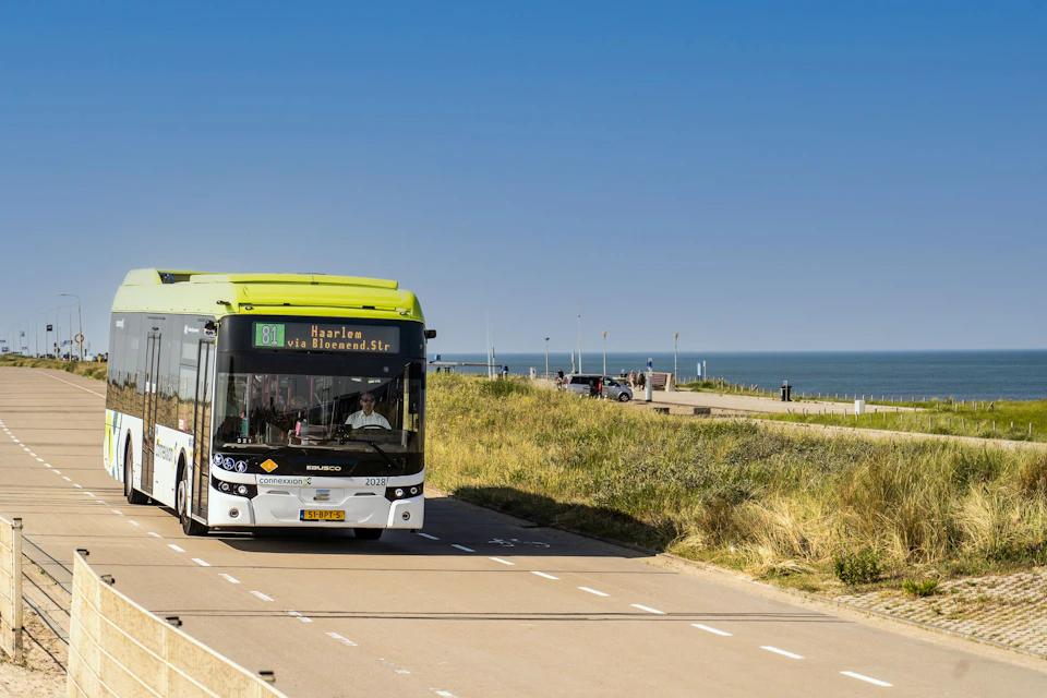 A public bus with route number 81 to Haarlem drives on a coastal road with grassy dunes and the sea in the background.