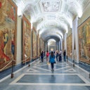 People walking through a museum hallway lined with large tapestries on both sides and a detailed, arched ceiling.
