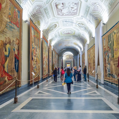 People walking through a museum hallway lined with large tapestries on both sides and a detailed, arched ceiling.