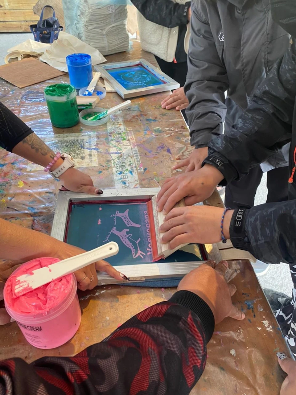 People working together on a screen printing project, using colorful inks on a wooden-framed screen on a table.