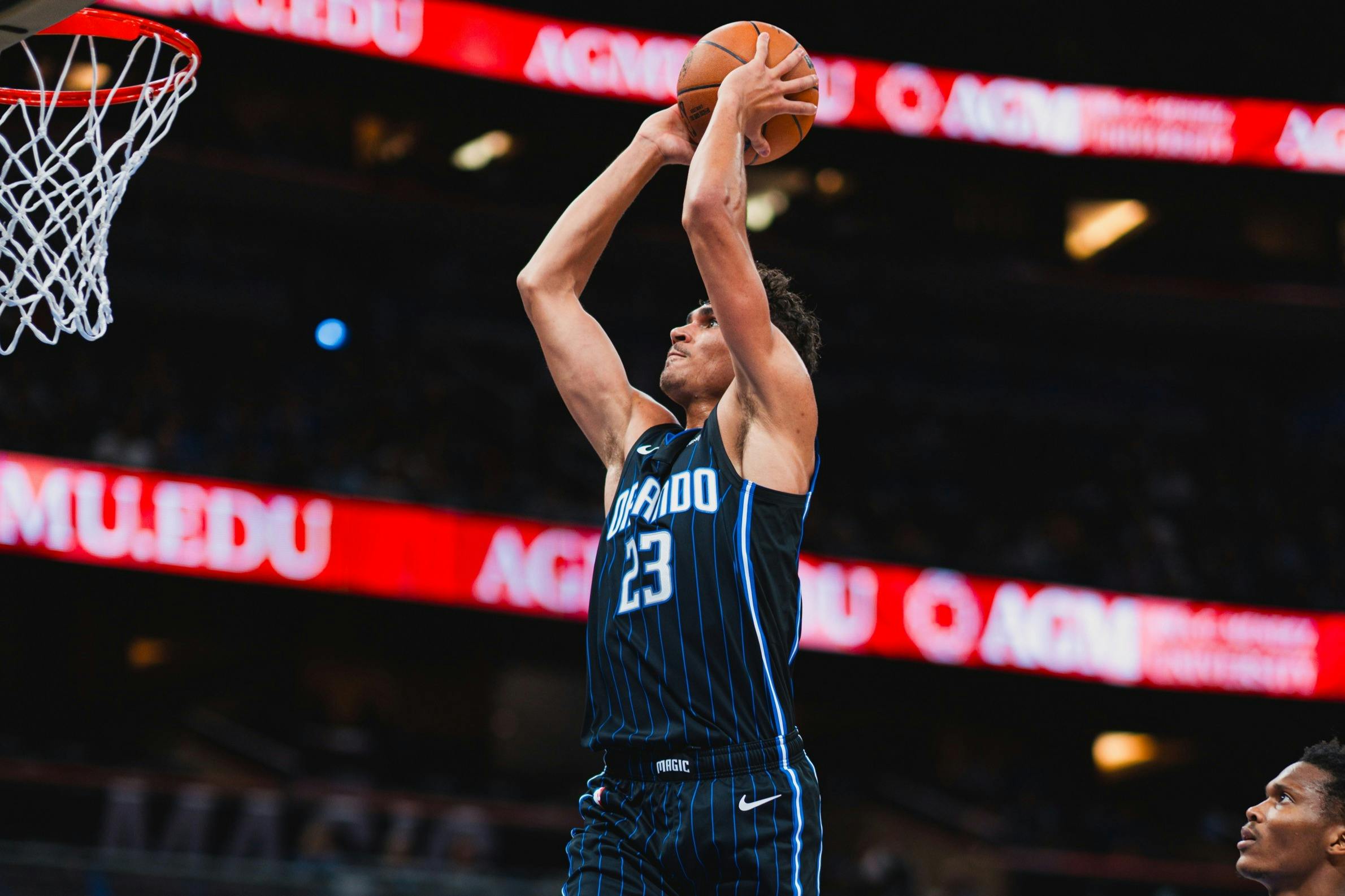 Ο μπασκετμπολίστας φορώντας μια μπλε στολή "Orlando" πηδάει για να σουτάρει την μπάλα σε μια αρένα με μια κόκκινη οθόνη LED στο παρασκήνιο.