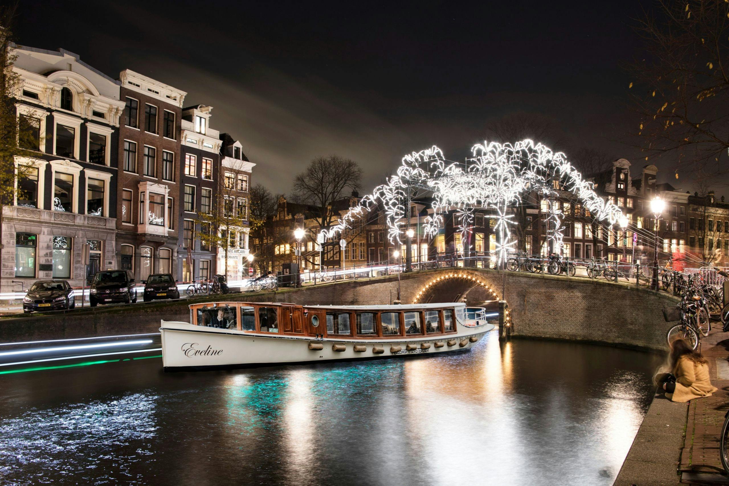 A boat named "Eveline" sails on a canal at night beneath a bridge adorned with festive white lights, surrounded by historic buildings.
