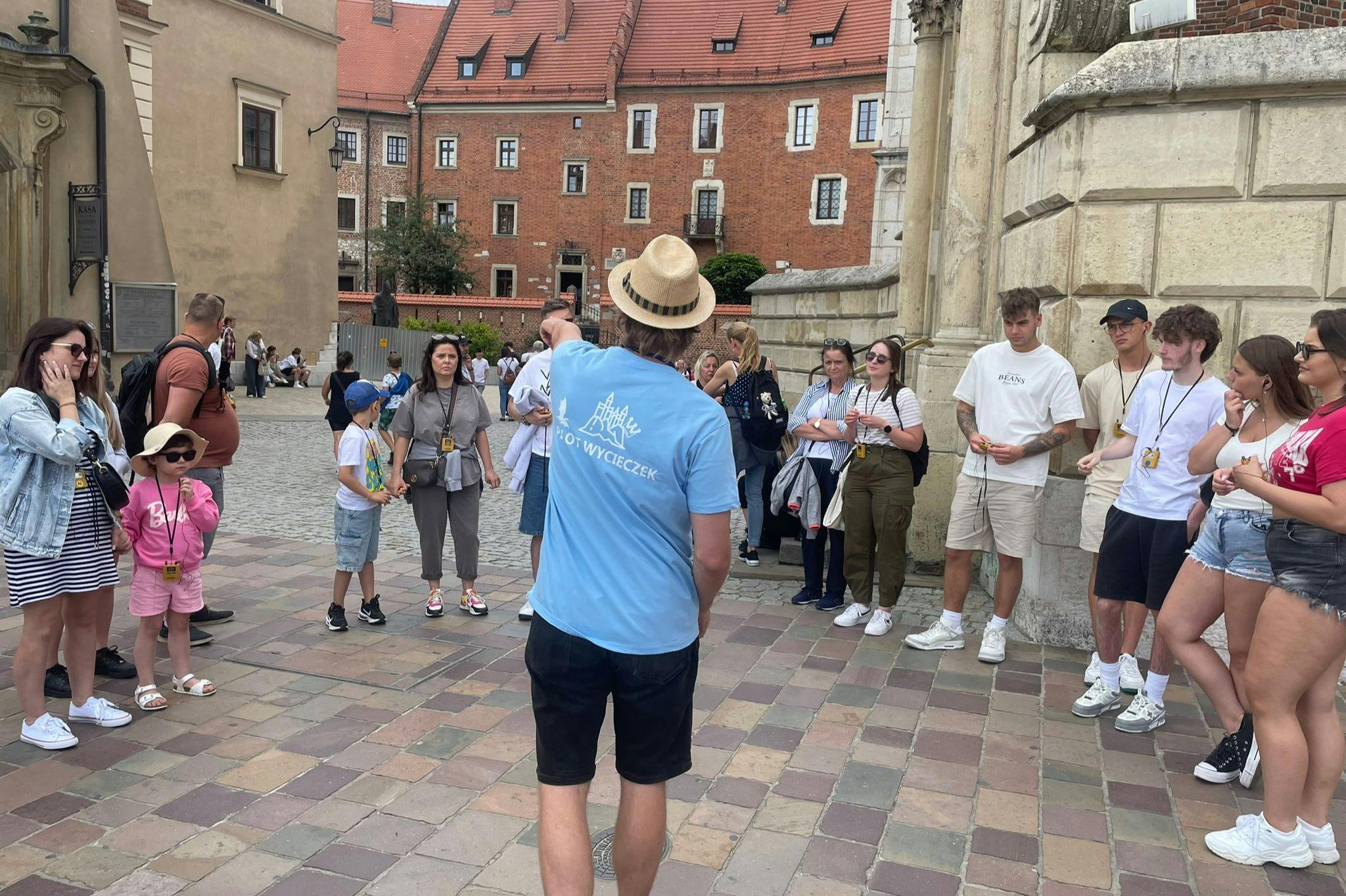 一群人站在鹅卵石广场上，有些人穿着蓝色衬衫和帽子，在历史建筑附近听导游讲解。