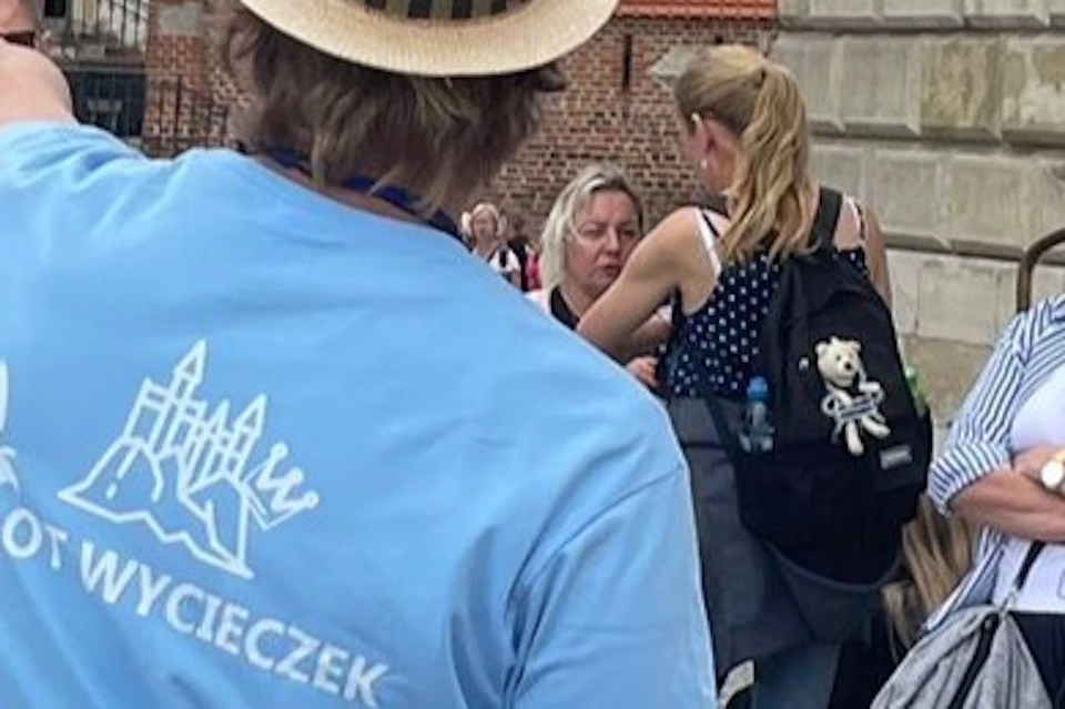 A group of people standing on a cobblestone square, some listening to a guide in a blue shirt and hat, near historic buildings.