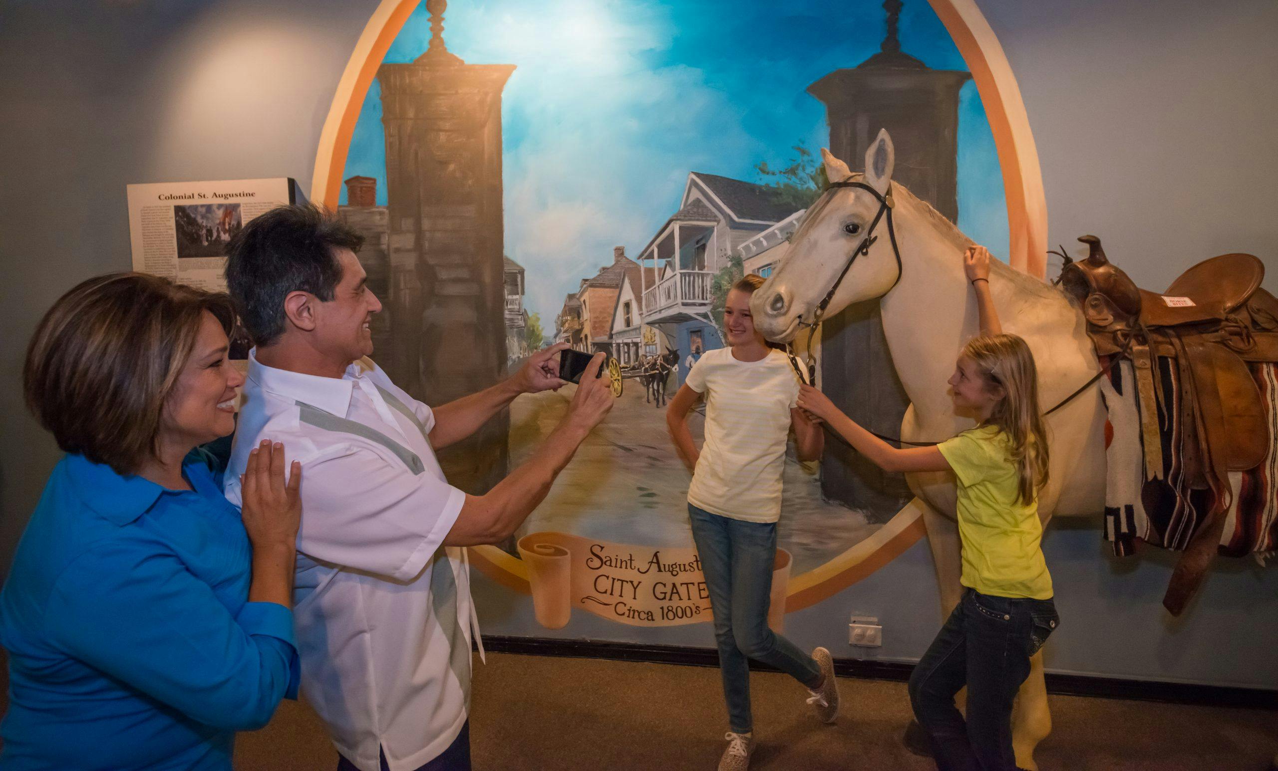 A smiling man takes a photo of a woman posing in front of a mural depicting Saint Augustine's historical city gate. Another woman stands beside him.