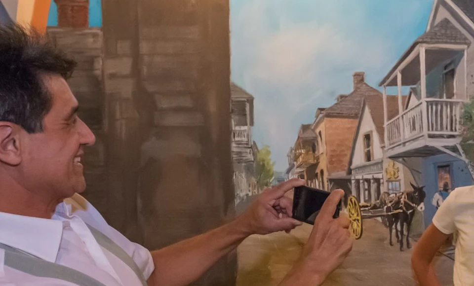 A smiling man takes a photo of a woman posing in front of a mural depicting Saint Augustine's historical city gate. Another woman stands beside him.