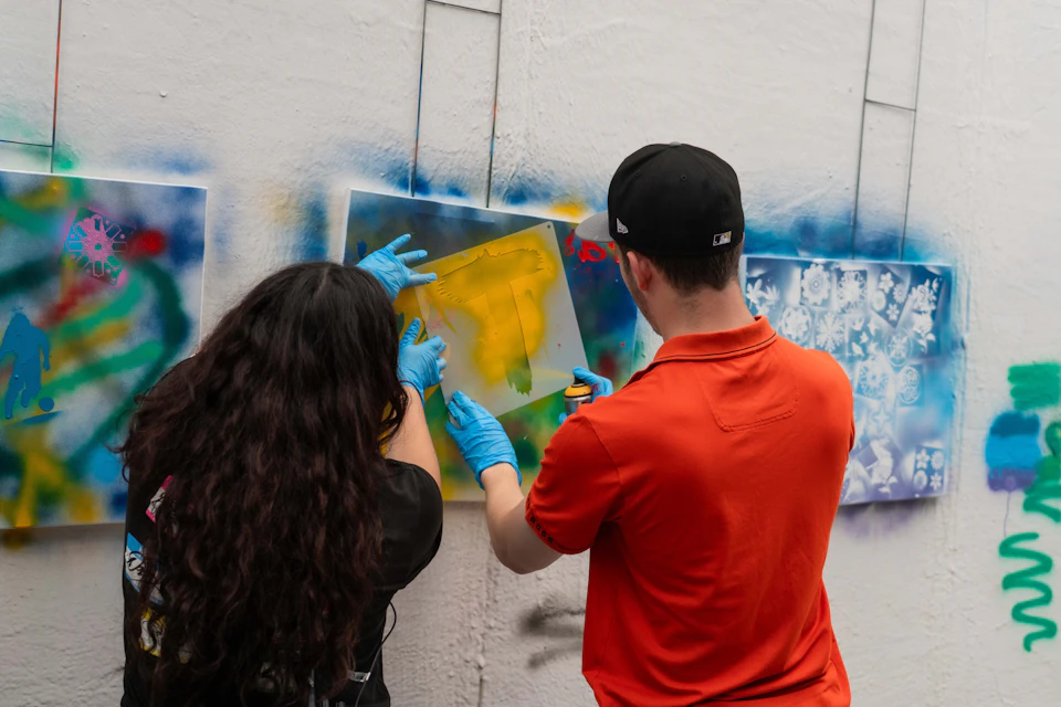 Two people wearing gloves and holding a stencil create graffiti art on an outdoor wall.